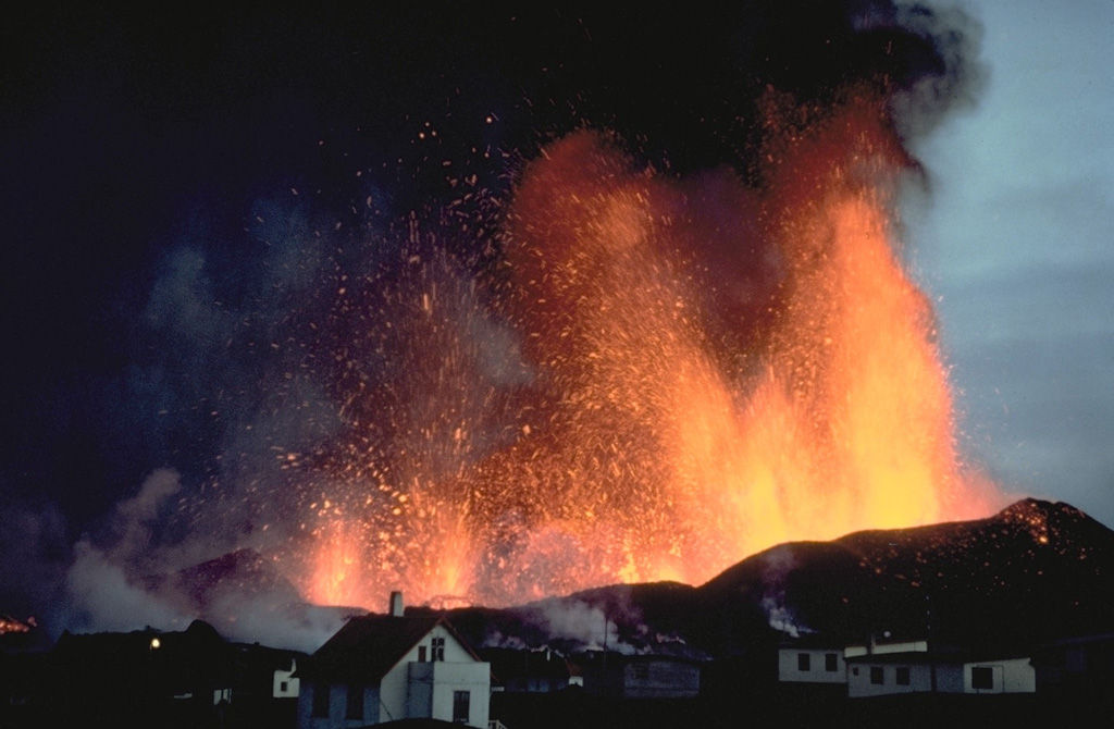 Fuentes de lava sobre la ciudad de Vestmannaeyjar en marzo de 1973. La erupción comenzó el 23 de enero de 1973, con eyecciones explosivas de una fisura parcialmente submarina con tendencia NNE-SSW que cortó el cono Helgafell. Las fuentes de lava construyeron el cono de cenizas de Eldfell y alimentaron flujos de lava que llegaron al mar. La tefra sepultó más de 100 casas y los flujos de lava inundaron partes de la ciudad y amenazaron su puerto. Se debaten los efectos de los enérgicos esfuerzos por frenar el avance de la lava rociándola con agua de mar fría, pero el flujo se estabilizó en un punto que mejoró el puerto natural. Foto con copyright de Katia y Maurice Krafft, 1973.