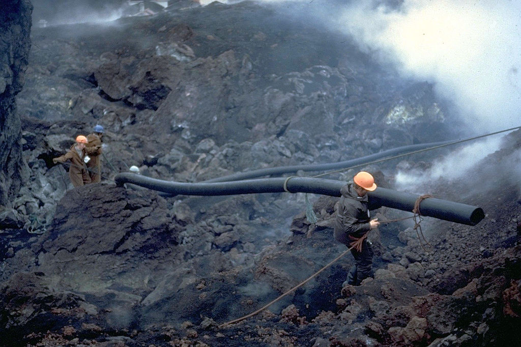Trabajadores transportan las tuberías utilizadas durante un intento masivo de enfriar el flujo de lava que avanza producido por la erupción del volcán Heimaey en 1973. El bombeo de agua comenzó el 6 de febrero, inicialmente con una sola bomba de gran tamaño, y luego con bombas más grandes enviadas por avión desde Islandia y Estados Unidos. El 1 de marzo llegó un buque de bombeo que roció agua desde el puerto. El enfriamiento local produjo barreras solidificadas que desviaron y detuvieron el flujo de lava. El flujo de lava creó nuevas tierras cuando entró en el mar y finalmente mejoró el único puerto de la isla. Foto con copyright de Katia y Maurice Krafft, 1973.