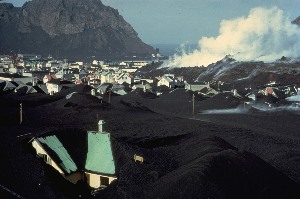 Una corriente de lava humeante avanza por la ciudad de Vestmannaeyjar en abril de 1973. Las casas en primer plano están parcialmente enterradas por los gruesos depósitos de ceniza del cono de ceniza de Eldfell, que comenzó a entrar en erupción el 23 de enero. El flujo de lava avanzó hacia el puerto de la ciudad, que se encuentra entre el flujo y el acantilado en la parte superior izquierda. El flujo cubrió un área de 300 m de ancho y 700 m de largo en la parte NE de la ciudad, enterrando 200 casas y edificios durante un período de dos semanas de extrusión a finales de marzo y principios de abril. Foto con copyright de Katia y Maurice Krafft, 1973.